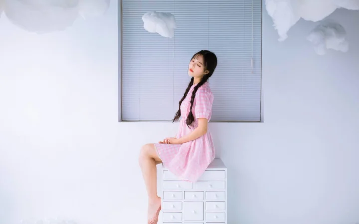 一名身穿粉色连衣裙、留着双辫的女性坐在白色抽屉柜上，背景是明亮的室内空间。