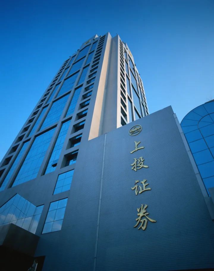 一座高层建筑从低角度向上延伸，立面在晴朗蓝天下呈现蓝灰色光影。