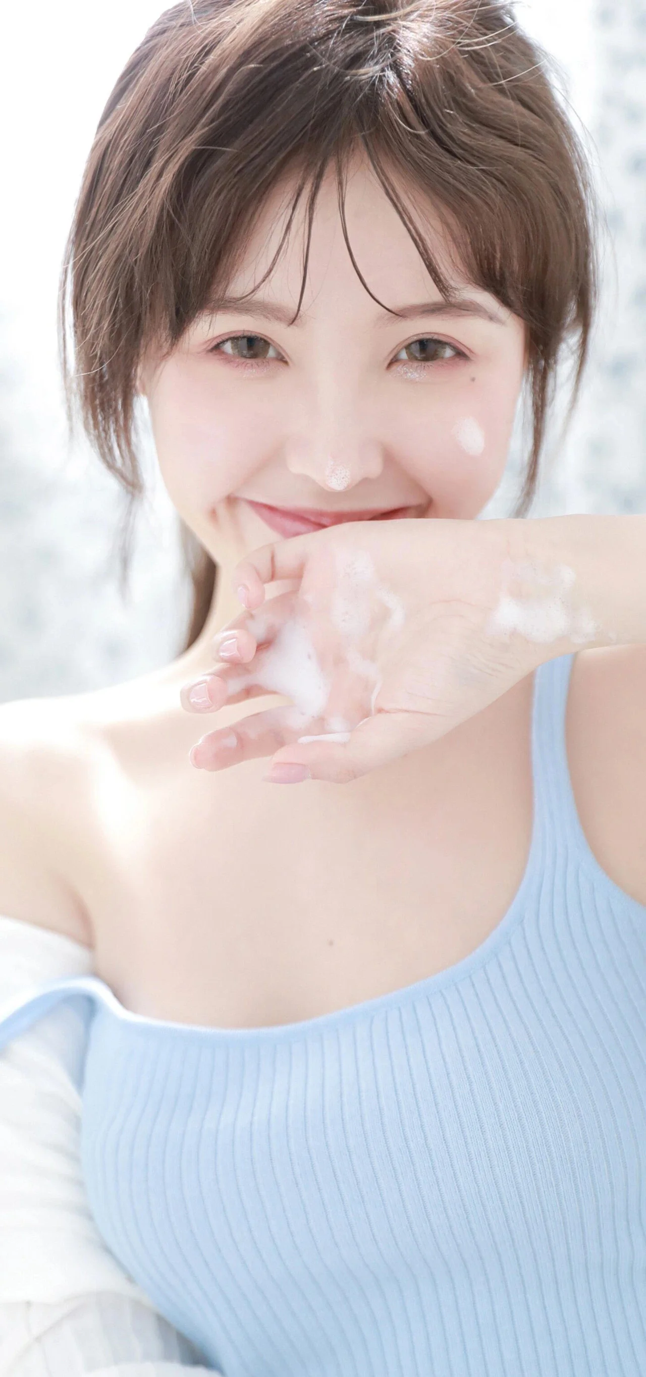 연한 파란색 상의를 입은 성인 여성이 손을 얼굴 앞에 대고 카메라를 향해 미소 짓고 있다.