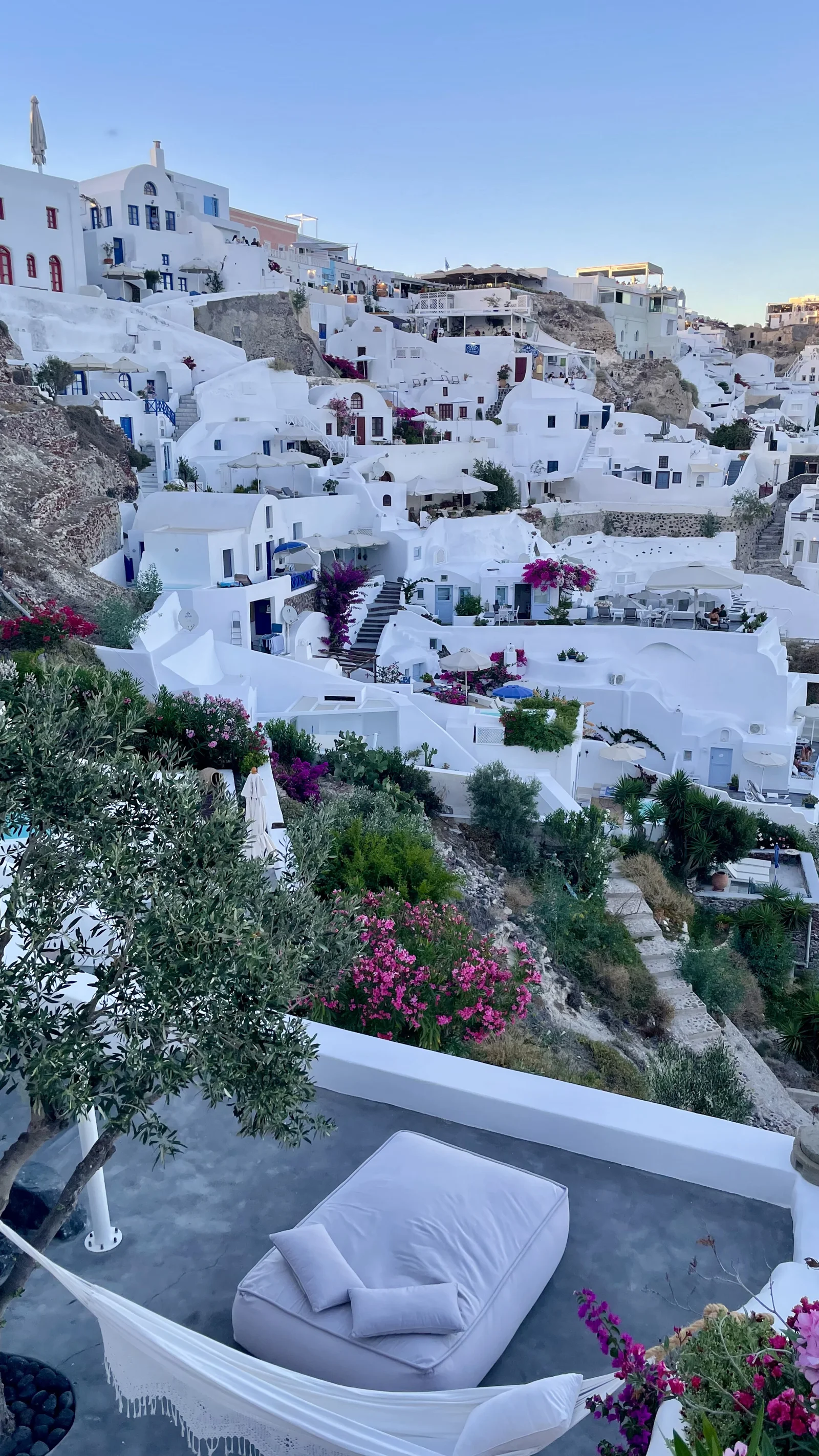 白色建筑沿山坡层叠分布，前景露台点缀着绿植和紫红色花朵。