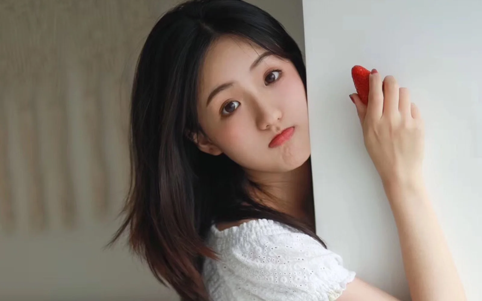 흰색 상의를 입은 검은 머리 여성이 딸기를 들고 벽 뒤에서 얼굴을 내밀고 있습니다.