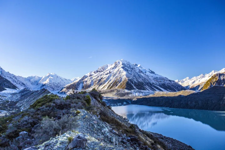 蓝天下的雪山环绕着宁静的蓝色湖泊，前景是 rocky 山坡与灌木。