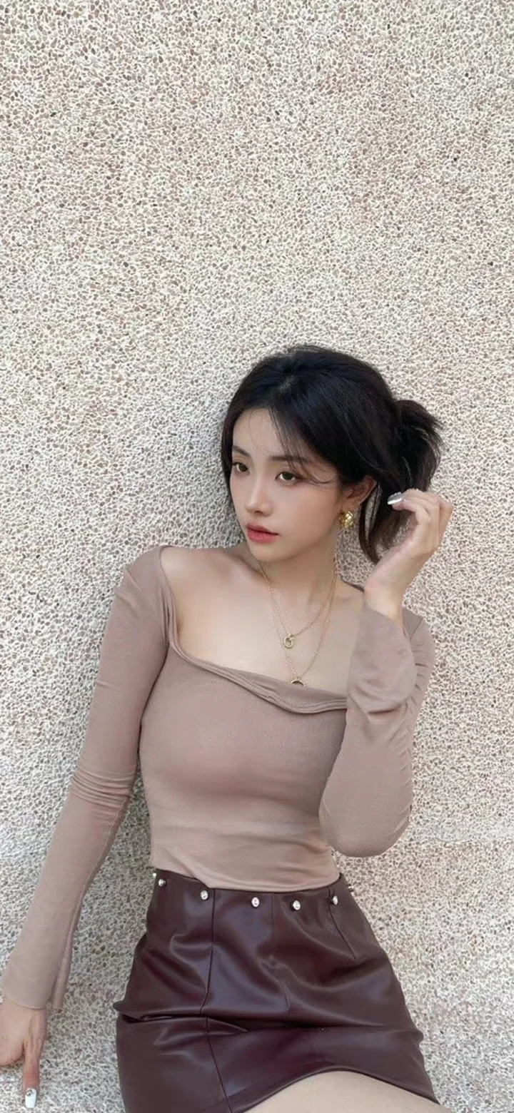 一位成年女性身穿浅棕色上衣和深色短裙，靠在纹理墙面前整理头发。