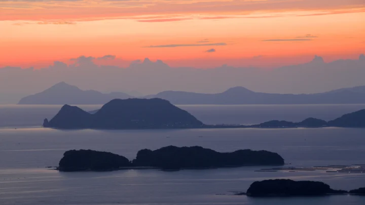 夕阳映照下的海面上分布着多座深色岛屿，远处可见层叠山峦与晚霞。