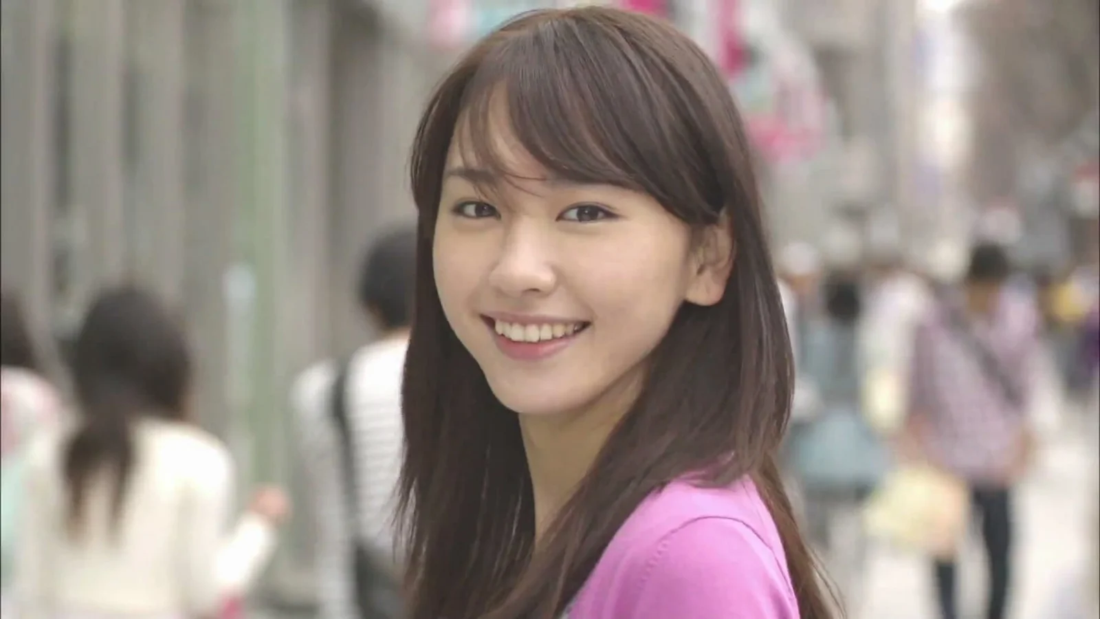 長い髪の女性がピンクのトップスを着て、ぼけた公共の場所でカメラに向かって微笑んでいる。