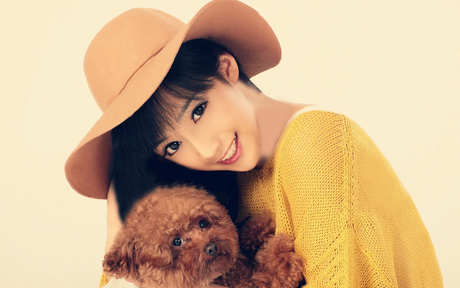 つばの広い帽子と黄色いニットを着た女性が、茶色の巻き毛の犬を抱えてカメラに微笑んでいる。