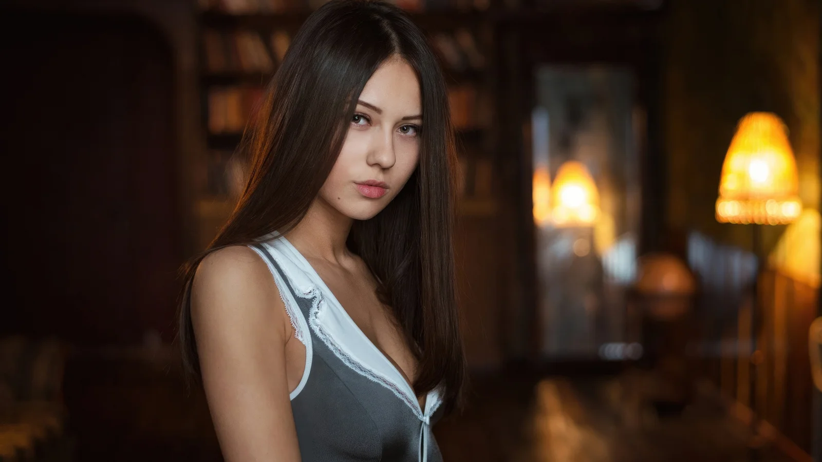 茶色の長い髪の女性が、暖かな薄暗い室内の照明を背景にグレーのノースリーブトップスを着て立っている。