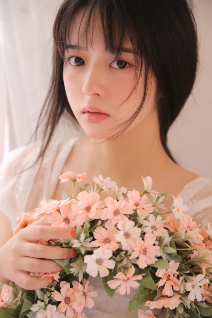 여성이 분홍색 꽃다발을 들고 부드러운 밝은 배경 앞에서 촬영된 클로즈업 인물 사진입니다.