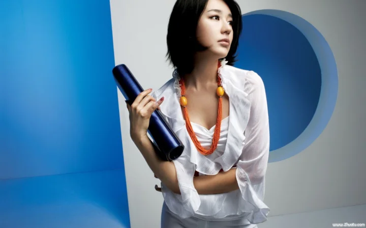 一名成年女性穿著白色上衣，手持深藍色圓筒物體站在藍白色幾何背景前。