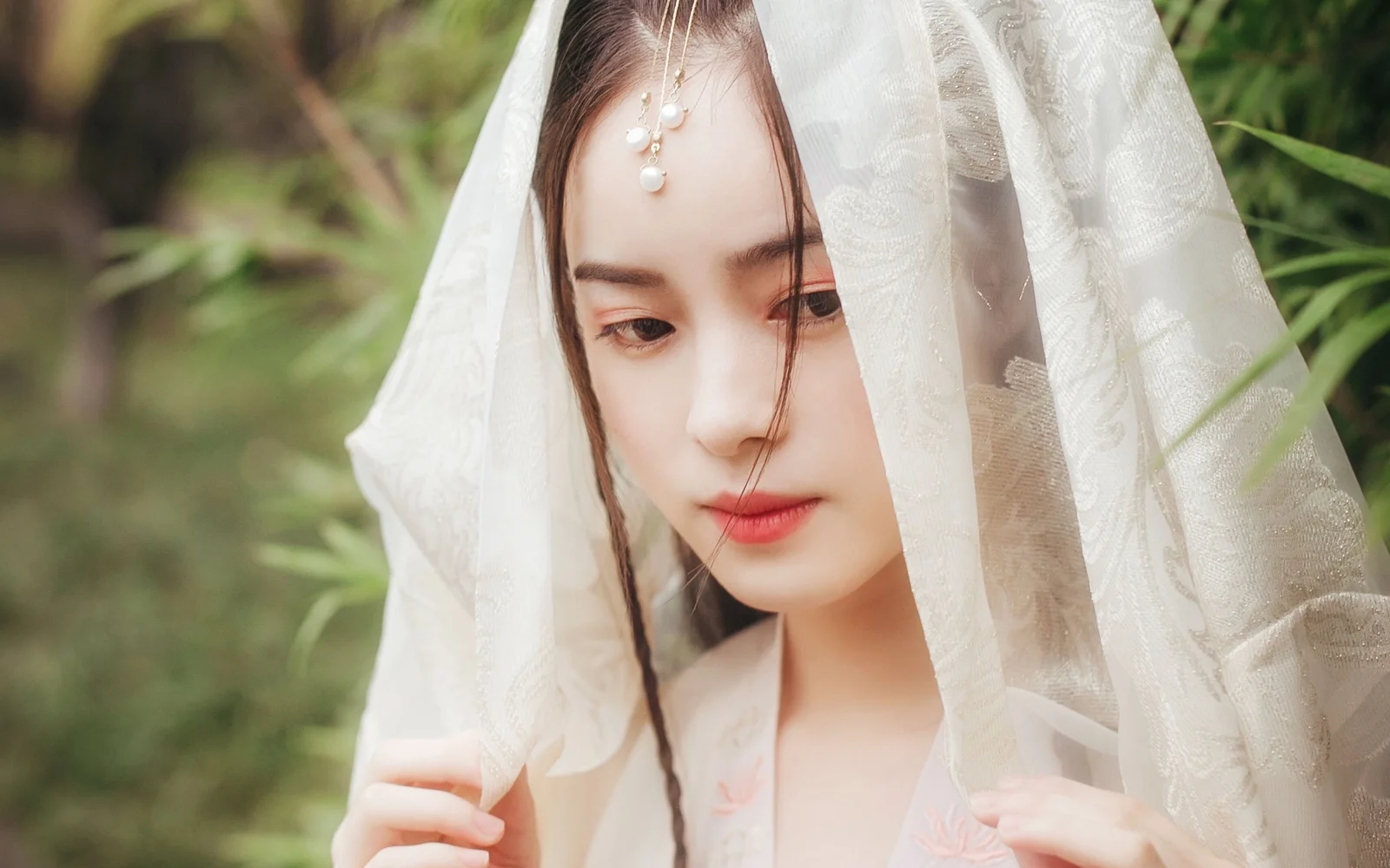 一位佩戴白色面纱和额饰的成年女性站在绿意背景前。