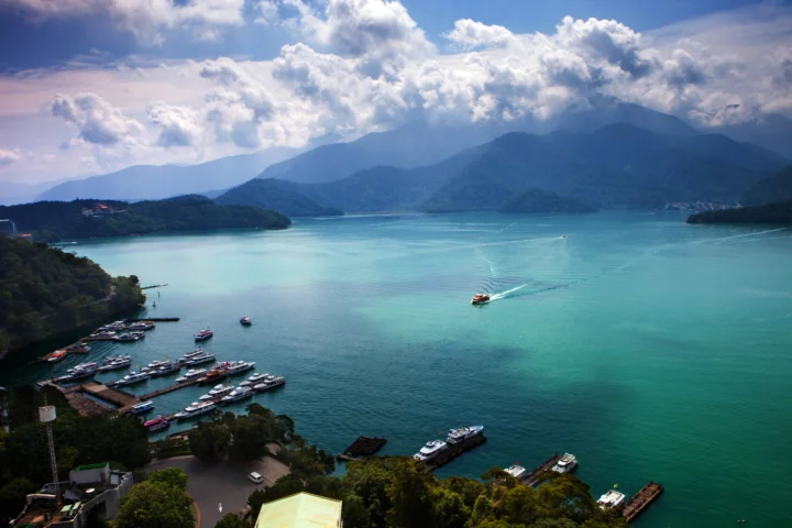 碧 turquoise 湖水延伸至群山之间，天空覆盖着层叠的白云，岸边可见停泊的小船。