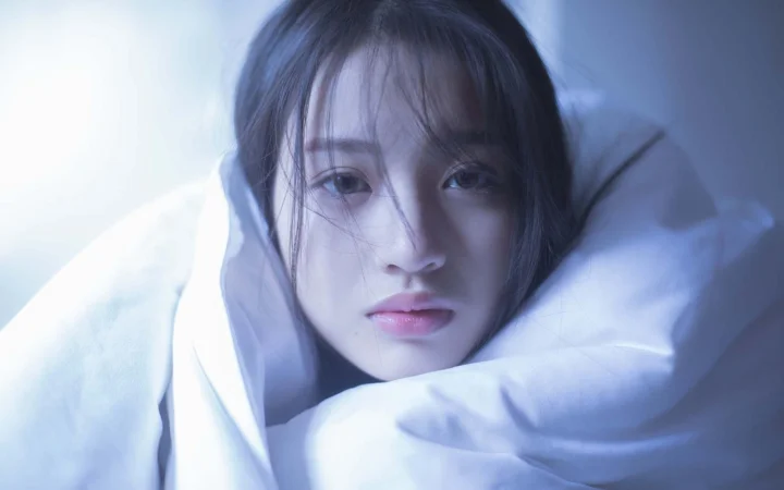 一名女性在冷蓝色光线下被白色床品包围，面向镜头呈现近距离肖像。