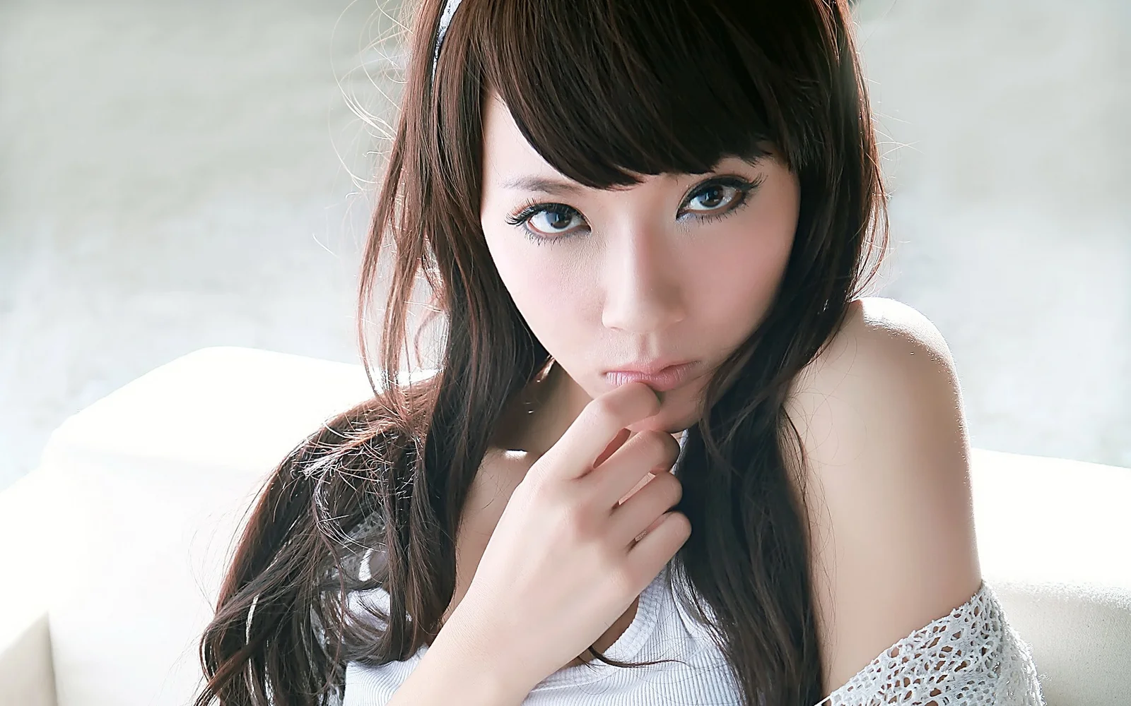 一位棕发女性坐在室内浅色沙发上，手指轻触下巴并注视镜头。