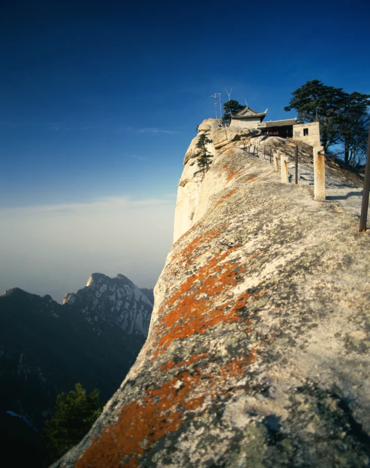 险峻的岩石山崖上坐落着一座古寺，远处群峰笼罩在湛蓝天空下。