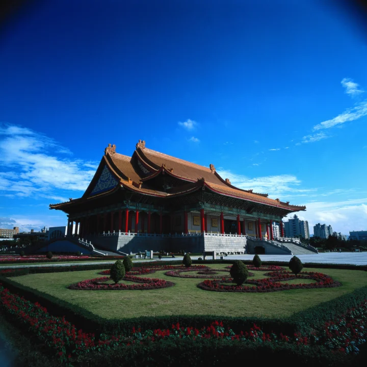 一座传统中式宫殿坐落在修剪整齐的花园中，背景是明亮的蓝天和城市建筑。