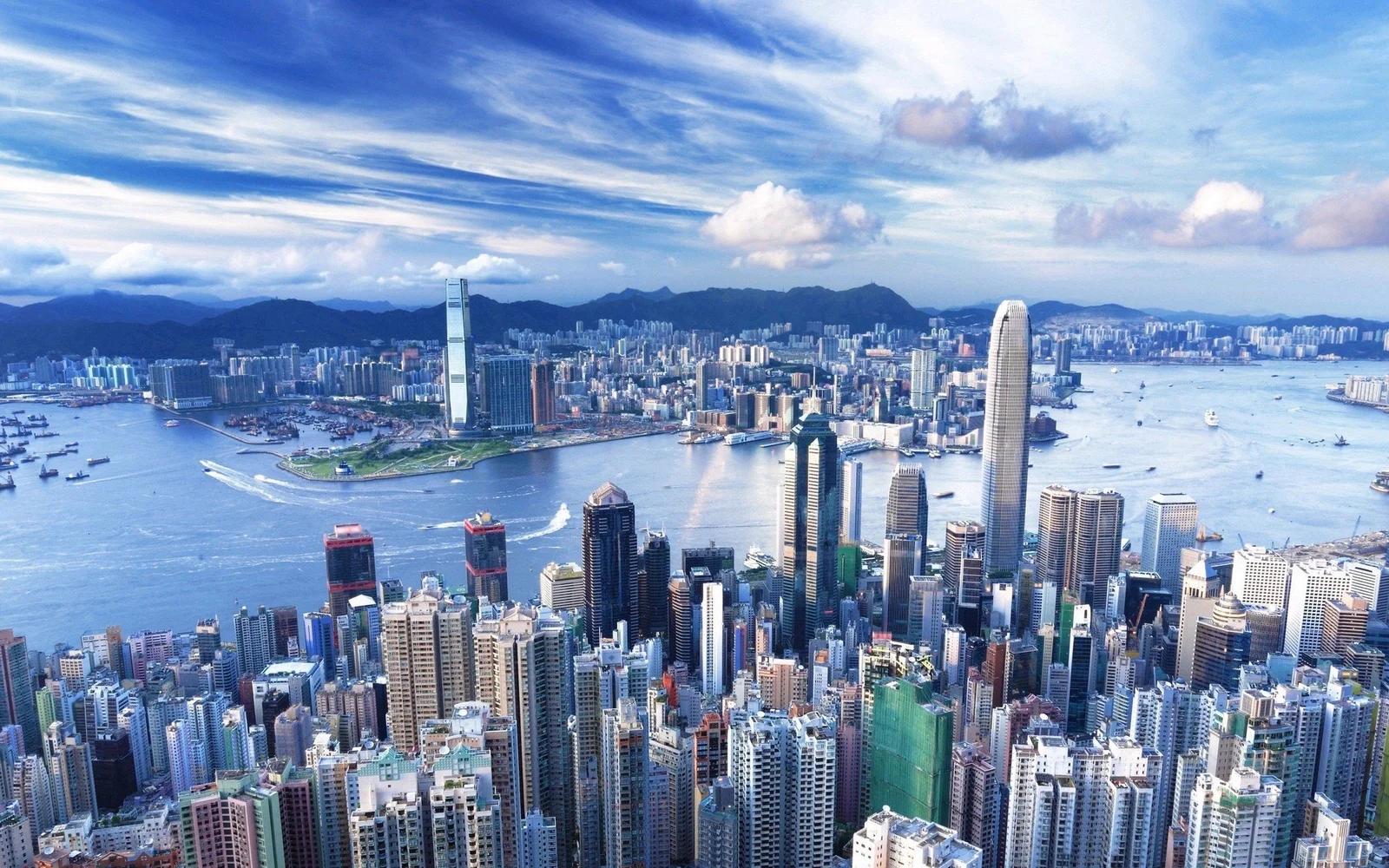 密集的滨海城市建筑环绕着繁忙的海湾，远处可见山脉和蓝天白云。