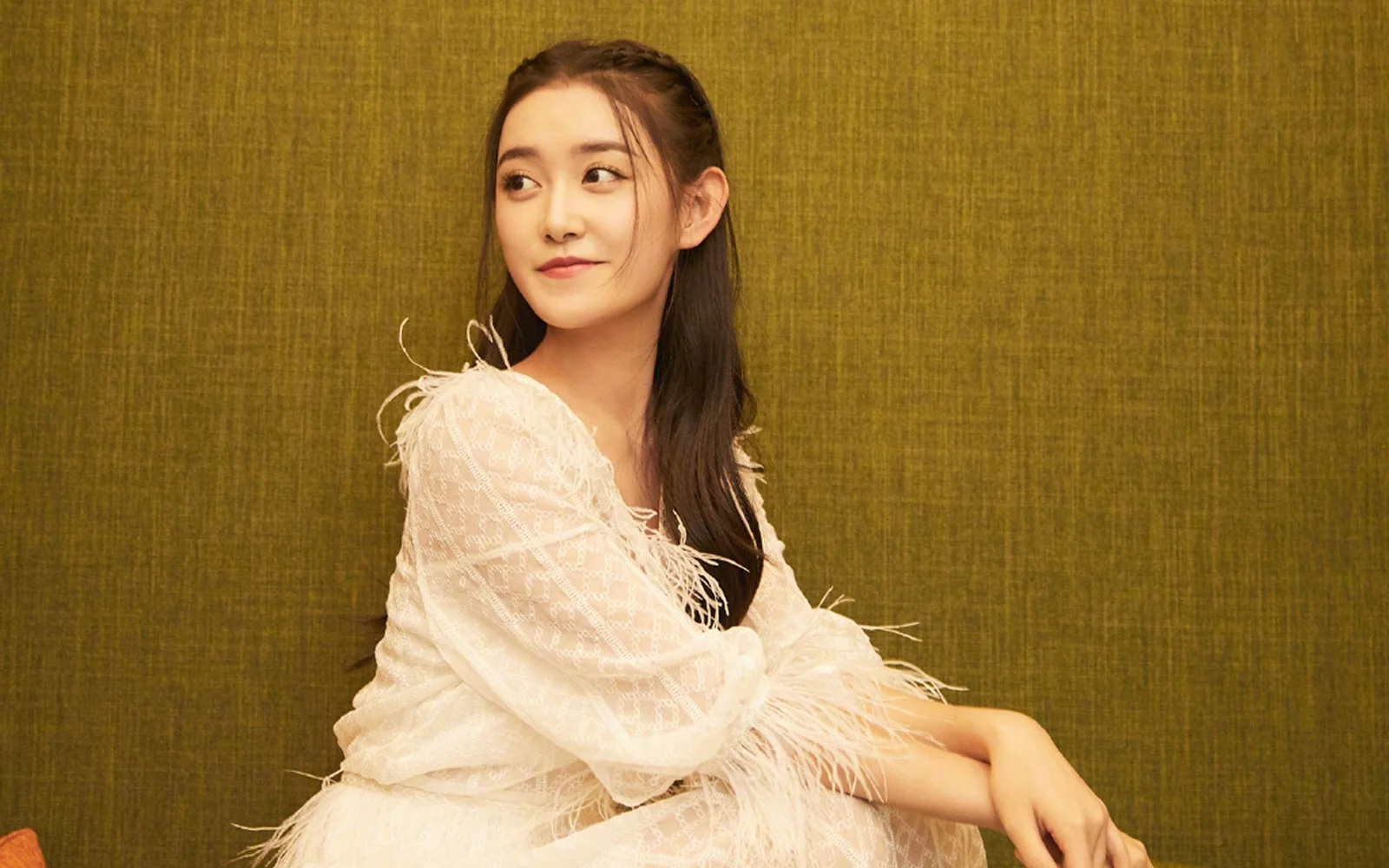 흰색 깃털 장식 드레스를 입은 성인 여성이 갈색 질감 배경 앞에 앉아 있습니다.