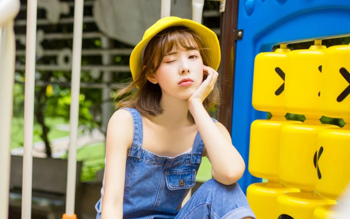 一名戴黄色帽子、穿牛仔背带裤的年轻女子在户外游乐设施旁托腮闭眼。