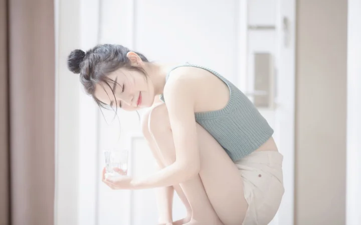 一名成年女性坐在明亮的室内窗边，手持玻璃杯并低头微笑。