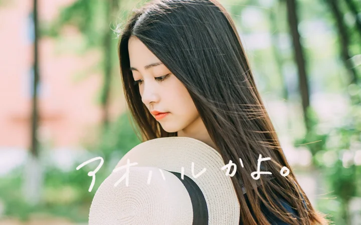長い髪の女性が麦わら帽子を手に森の中でうつむき加減に横顔を見せ、背景には柔らかな緑のボケが広がっています。