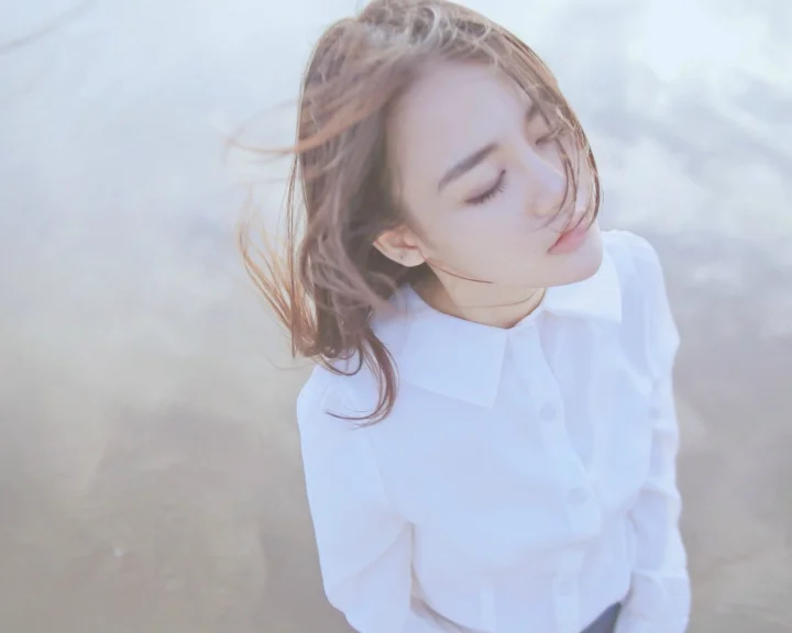 一名闭着眼睛的成年女子穿着白色衬衫站在浅色背景前。