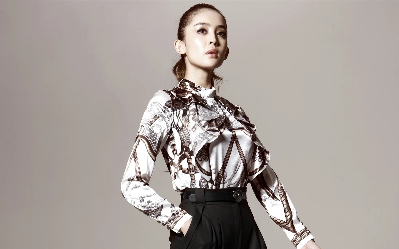 인쇄무늬 하이넥 블라우스를 입은 성인 여성이 회색 스튜디오 배경 앞에 서 있다.