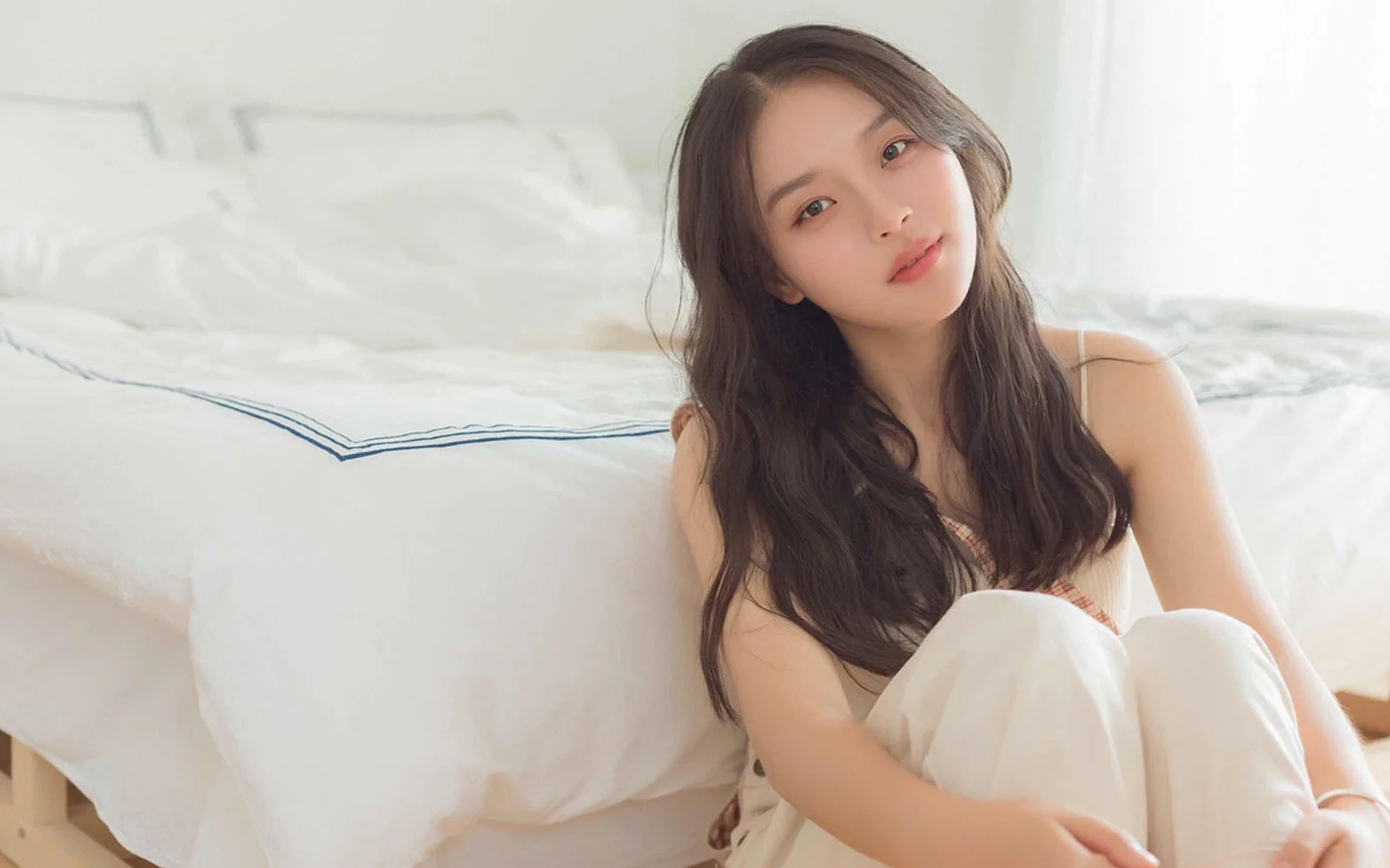 一名成年女性坐在明亮臥室的床上，面向鏡頭微笑。