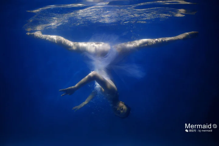 人魚の尾を身につけた人物が、深い青色の水中で水面の反射を受けながら漂っている。