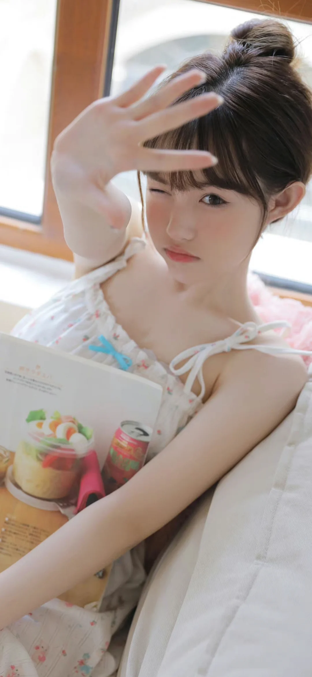 一名盘发女子在窗边举手遮挡阳光并眨眼，手中拿着一本打开的书或杂志。