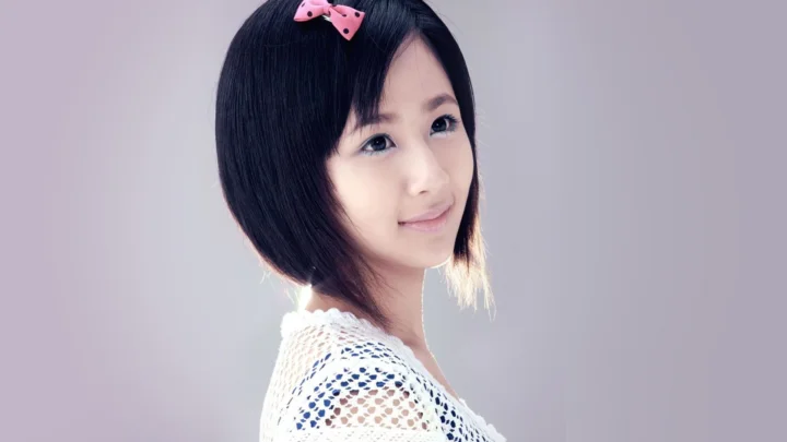 분홍색 리본 머리 장식을 한 검은 머리 여성이 밝은 배경 앞에서 옆모습으로 미소 짓고 있습니다.