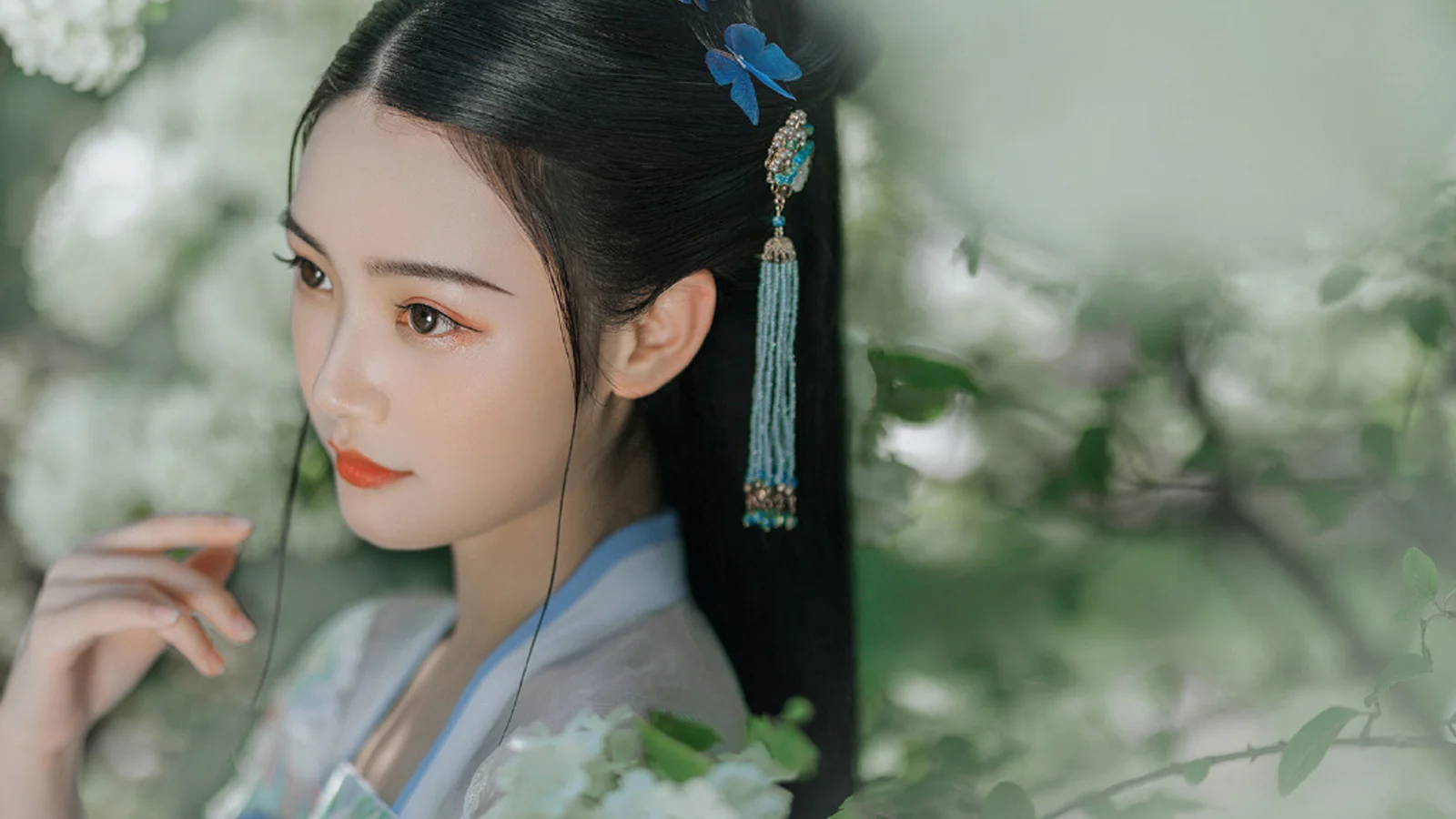 전통 의상을 입고 파란 머리 장식을 한 여성이 부드럽게 흐려진 푸른 잎사귀 사이에 서 있다.