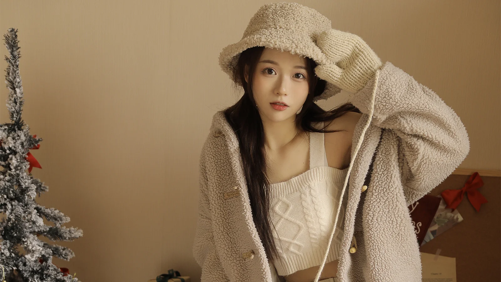 복슬복슬한 모자와 베이지색 겨울옷을 입은 성인 여성이 장식된 크리스마스 트리 옆에 서 있습니다.