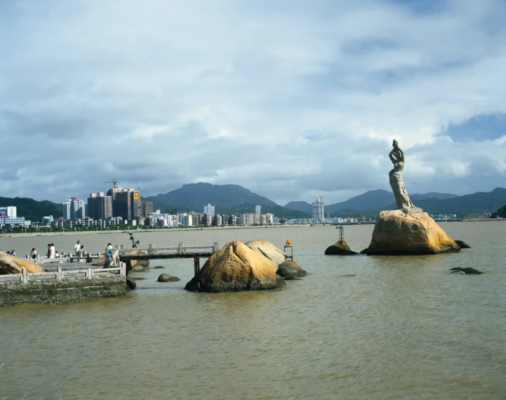 宽阔水面前是一座海滨城市，远处有群山和矗立在巨石上的雕像。