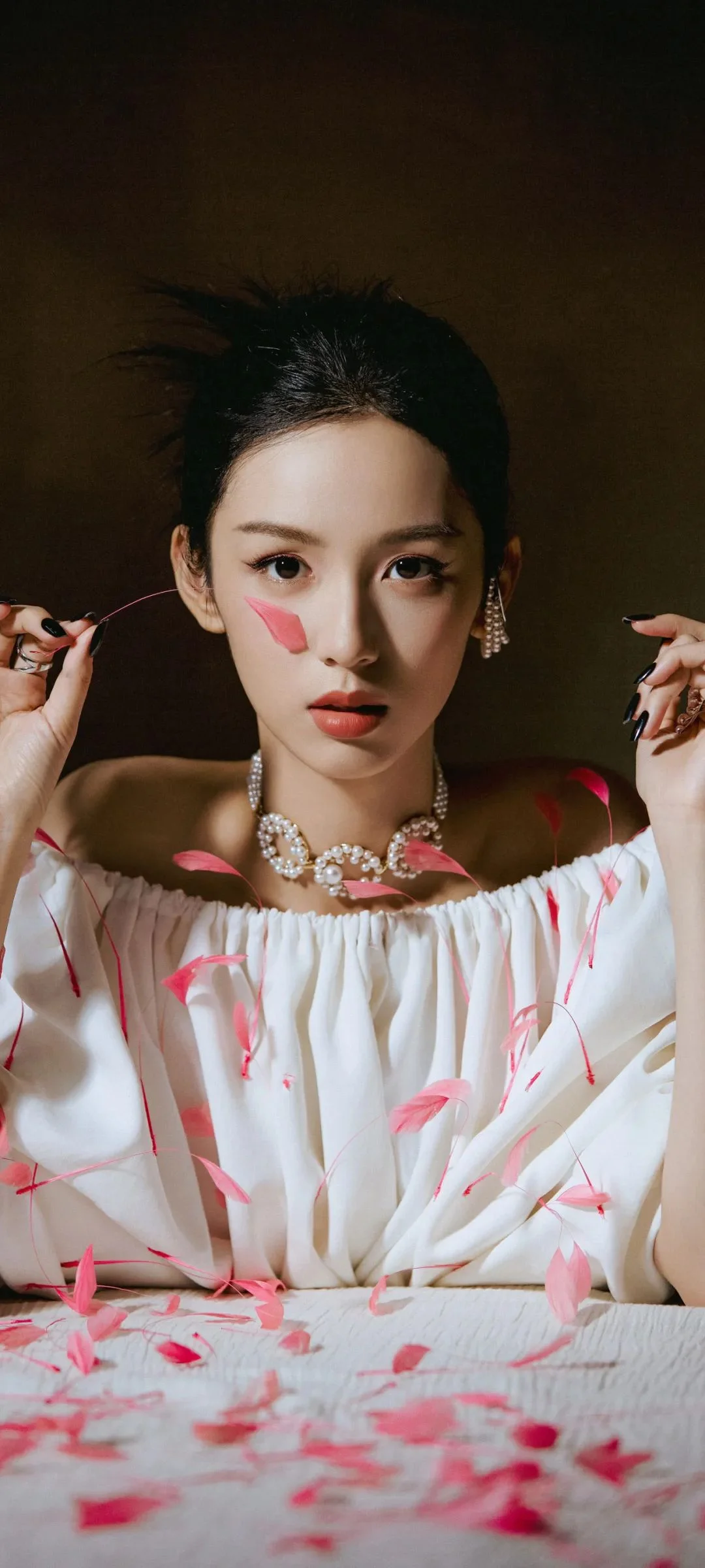 一位成年女性身穿白色露肩服装，手持细线，周围散落着粉色花瓣并佩戴华丽项链。