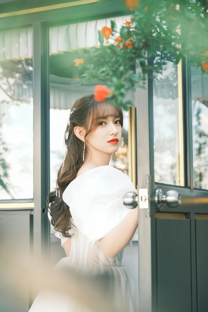 흰색 옷을 입은 성인 여성이 초록 잎과 주황색 꽃이 보이는 유리문 옆에 서 있다.
