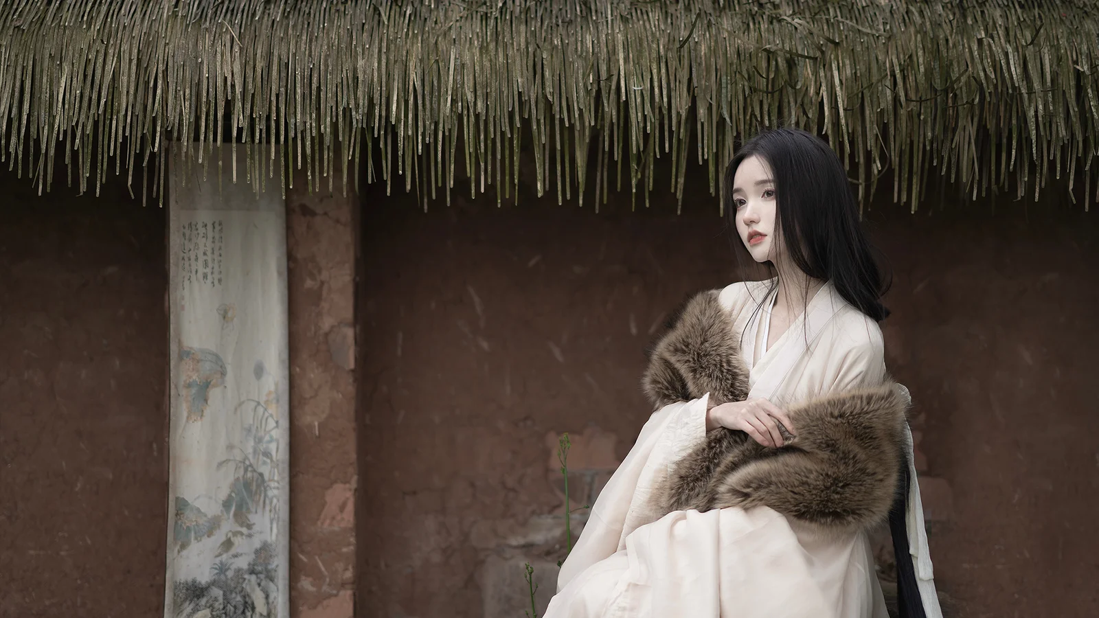 一名身穿浅色传统服饰并披着毛皮披肩的女子坐在茅草屋檐下。