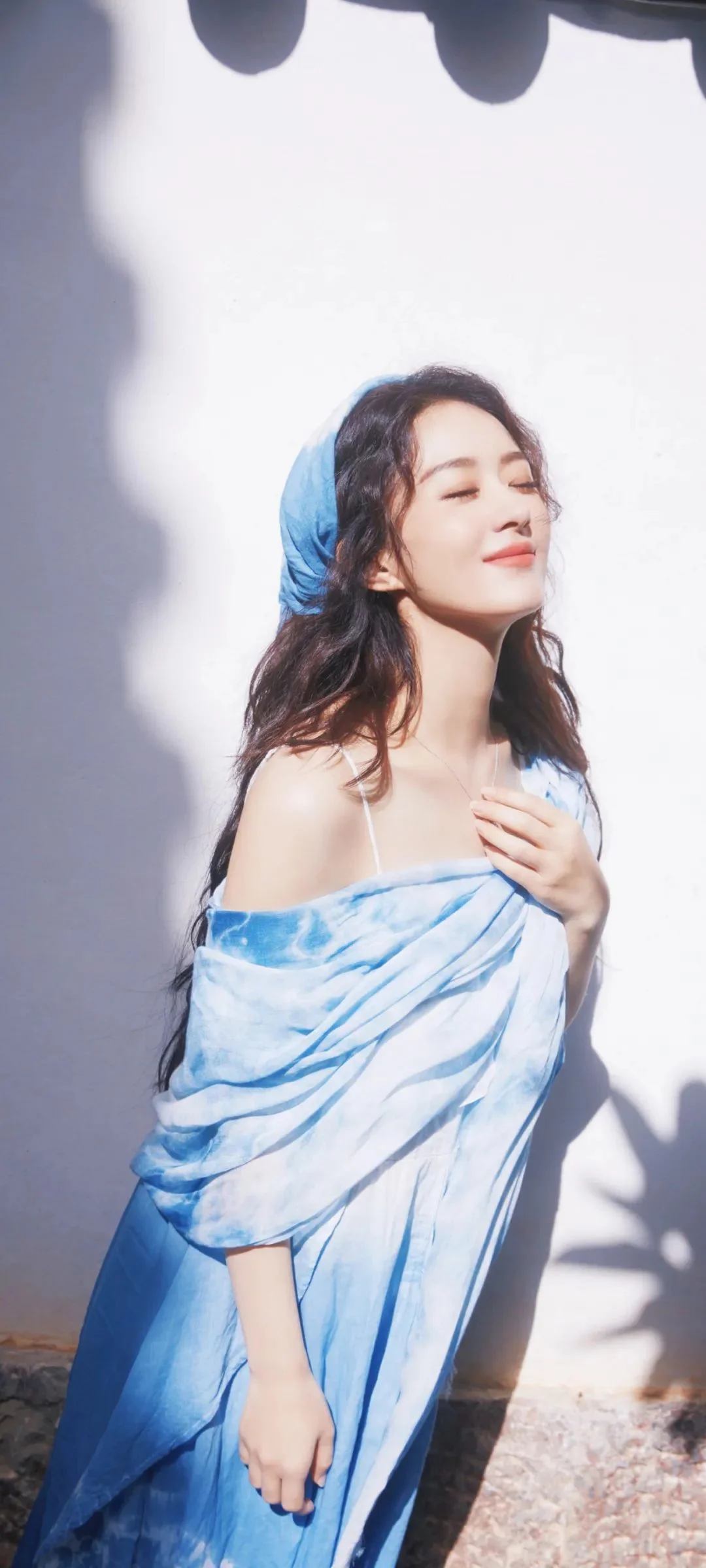 一位成年女性身穿蓝白色露肩裙，在明亮墙面前闭眼站立。