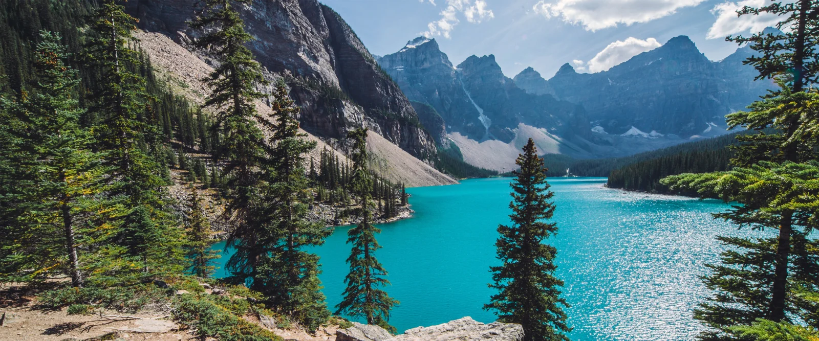 碧蓝湖泊 surrounded by evergreen forests and rugged mountains under a bright sky.