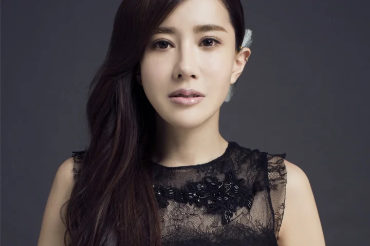 긴 머리의 여성이 검은 레이스 상의를 입고 짙은 회색 배경 앞에서 카메라를 바라보고 있습니다.