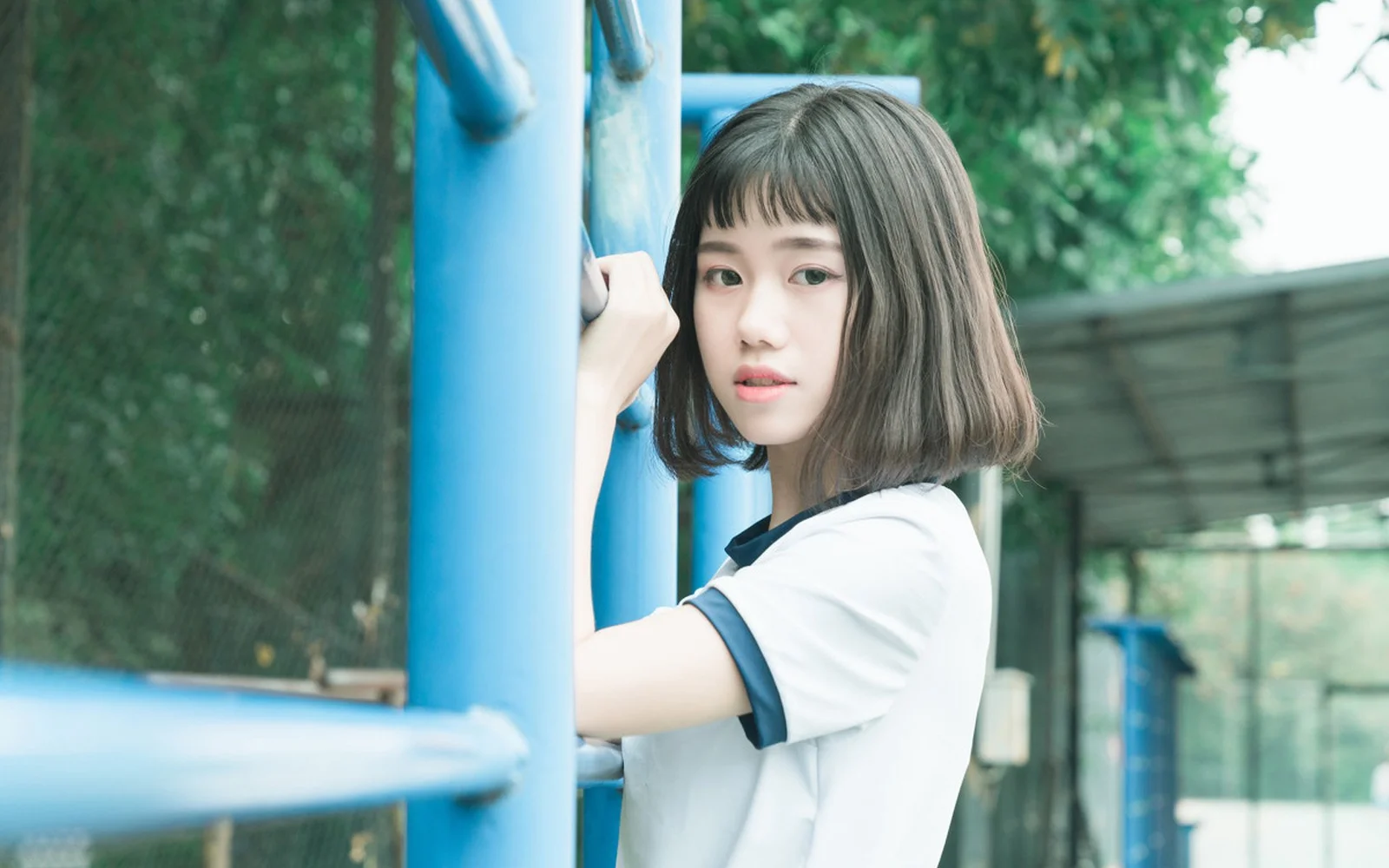 一名短发女性站在户外蓝色栏杆旁，背景是绿树和围栏。