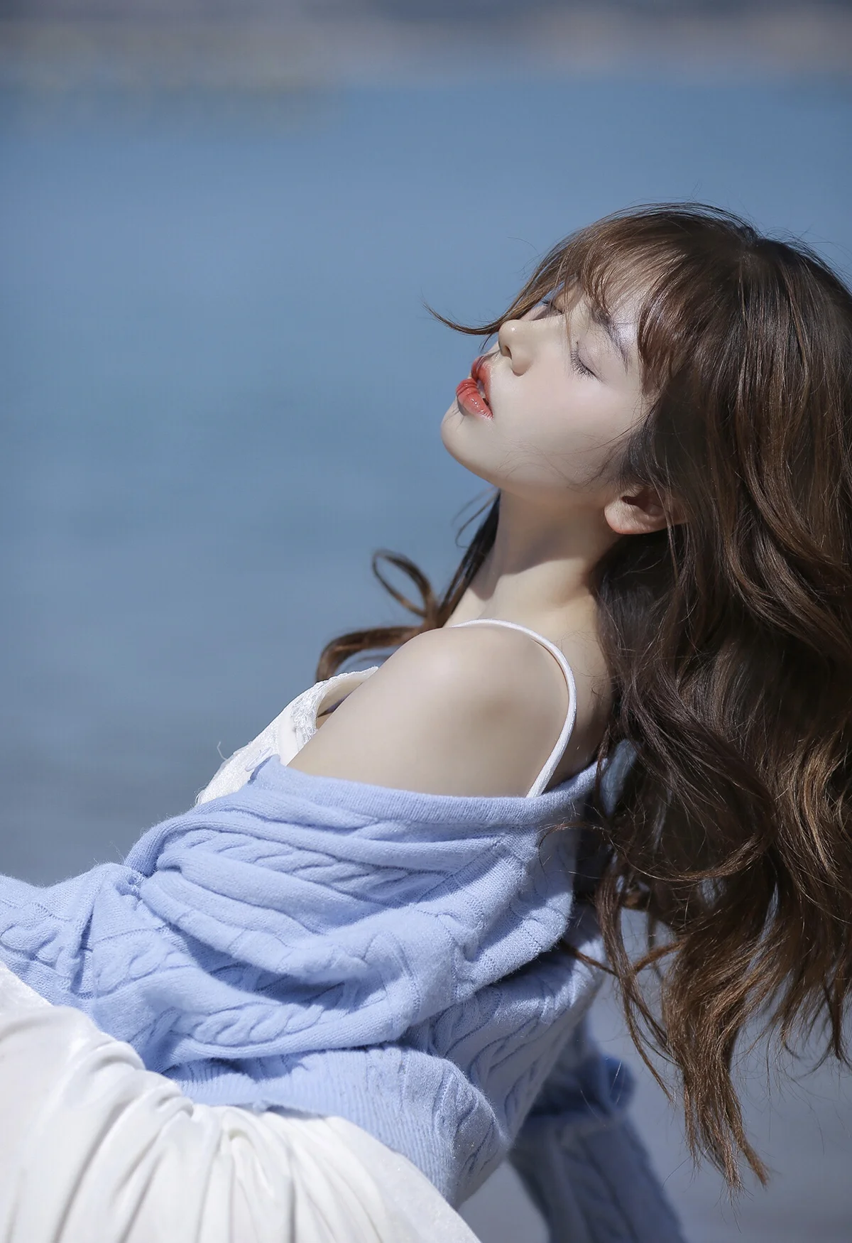 긴 머리의 여성이 눈을 감고 고개를 뒤로 젖힌 채 연한 파란색 담요를 두르고 파란 배경 앞에 앉아 있다.