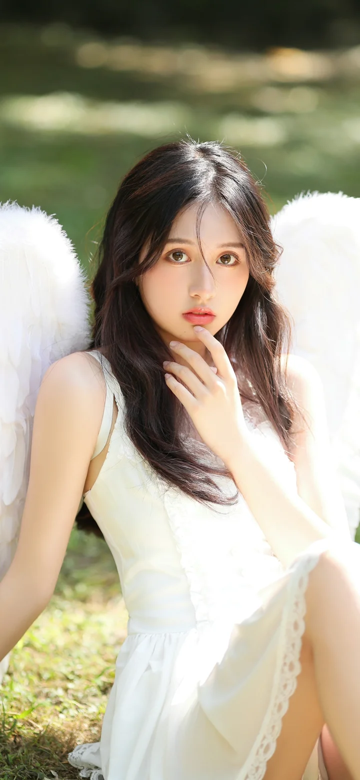 一名长发女子身穿白裙，背着白色翅膀坐在户外草地上。