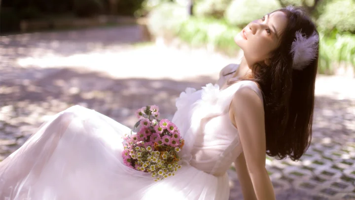 一名身穿白色连衣裙的女子坐在户外石板路旁，手捧一束鲜花并仰望前方。