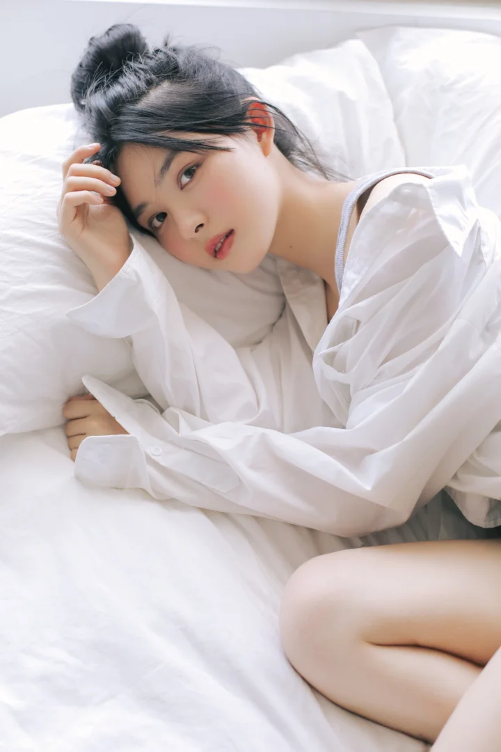 一名成年女子穿着宽松白衬衫侧卧在白色床铺上。