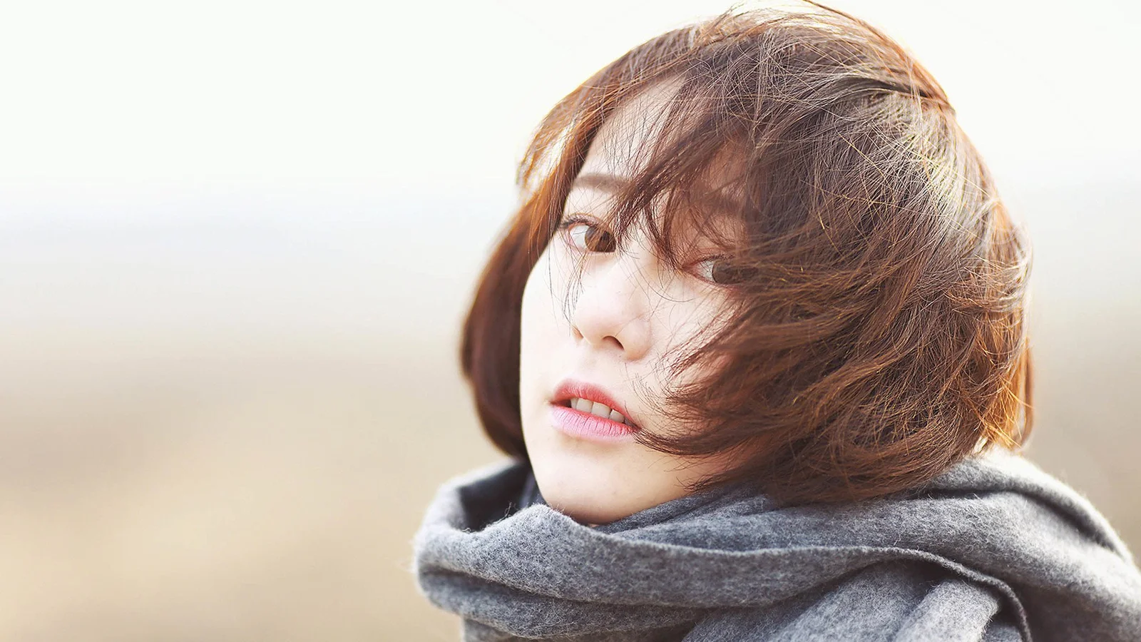 짧은 머리의 여성이 회색 목도리를 두르고 부드럽게 흐려진 야외 배경 앞에서 카메라를 바라보고 있습니다.