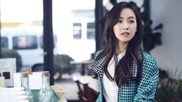 긴 머리의 여성이 청록색 무늬 재킷과 흰색 상의를 입고 실내 창가에 앉아 있습니다.