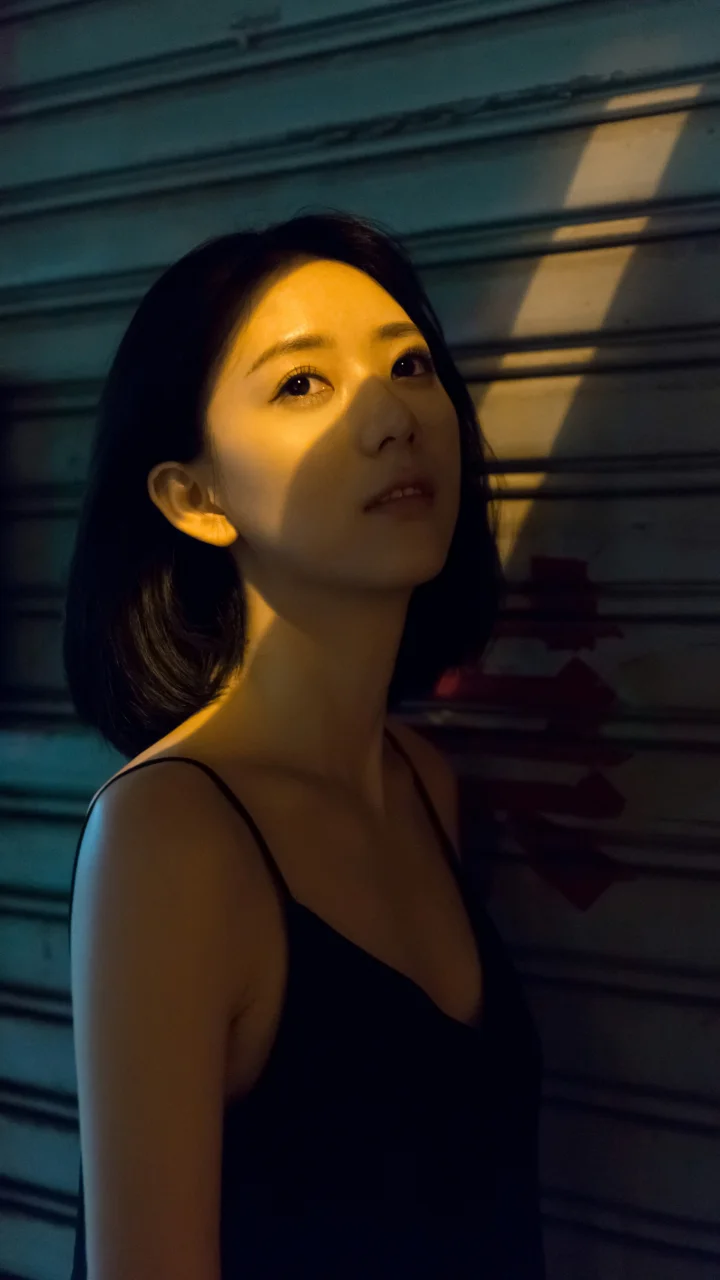 검은색 민소매 상의를 입은 성인 여성이 금속 셔터 앞에 서 있고 얼굴이 좁은 따뜻한 빛에 비춰지고 있다.