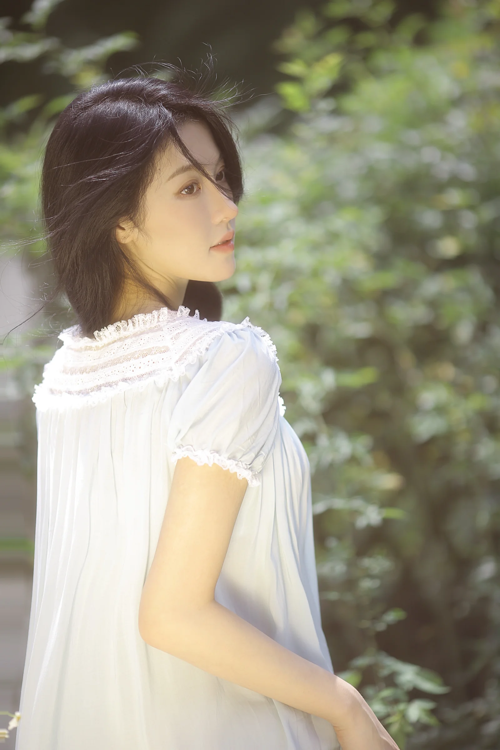 白いワンピースを着た女性が、緑の多い屋外で横顔を見せています。