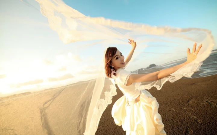 白いドレスを着た成人女性が、陽光に照らされた浜辺で両腕を広げています。