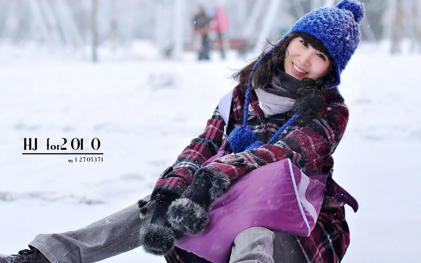 青いニット帽とチェック柄のコートを着た女性が雪の中に座って微笑んでいる。